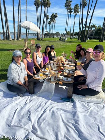 Group of friends enjoying a sunny bayfront brunch picnic under tall palm trees, low table with charcuterie and drinks, grassy waterfront and bridge in the background.