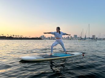 Person holding a yoga warrior pose on a stand-up paddleboard at sunset, calm marina waters with sailboats, palm trees and a city skyline in the background.