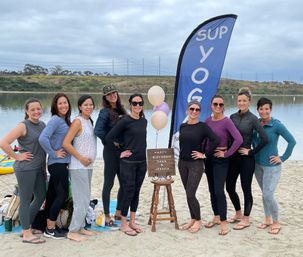 Smiling group of women posing on a lakeside sandy beach next to a tall SUP yoga banner, pastel balloons and a small birthday sign, with calm water and rolling hills in the background — outdoor paddleboard yoga birthday gathering.