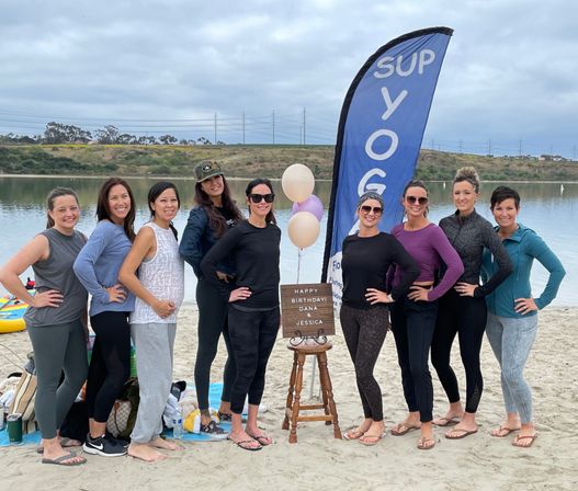 Smiling group of women posing on a lakeside sandy beach next to a tall SUP yoga banner, pastel balloons and a small birthday sign, with calm water and rolling hills in the background — outdoor paddleboard yoga birthday gathering.
