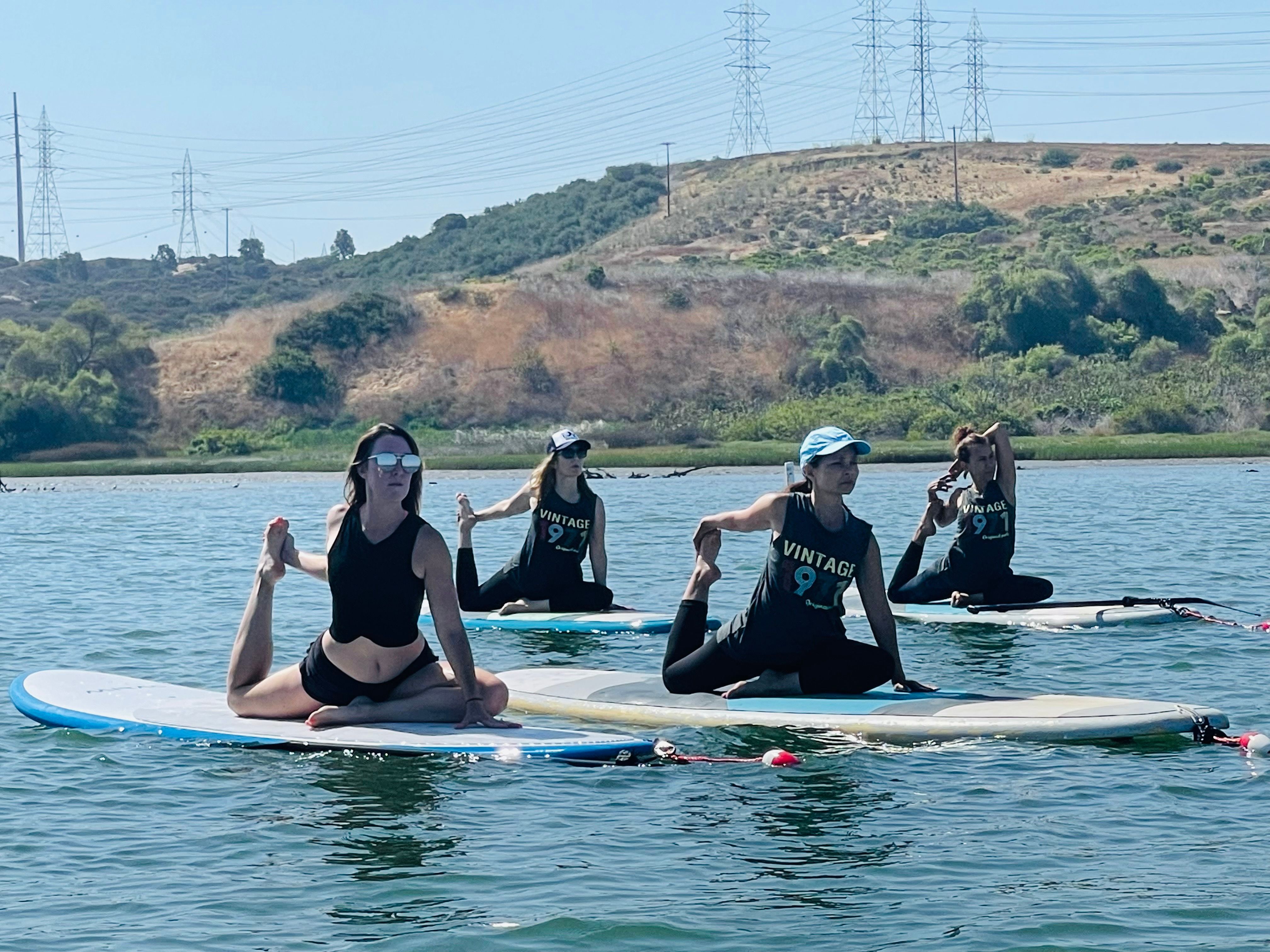 Four women practicing SUP yoga on paddleboards on a calm lake by sunlit hills, holding seated and quad-stretch poses for outdoor paddleboard fitness