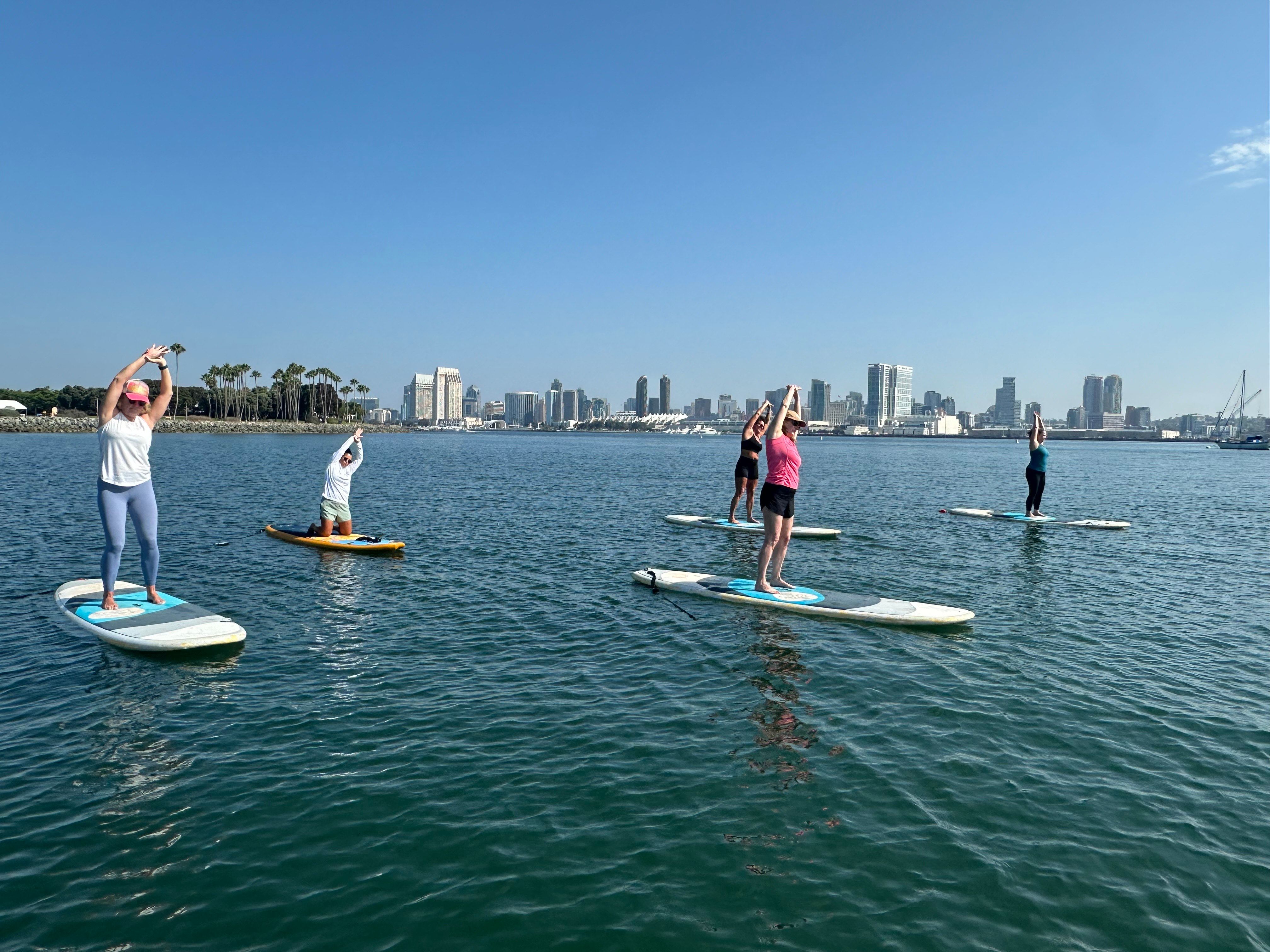 Five paddleboarders doing yoga stretches on a calm bay under a sunny blue sky with palm trees and an urban skyline in the background