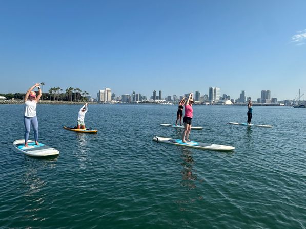 Five paddleboarders doing yoga stretches on a calm bay under a sunny blue sky with palm trees and an urban skyline in the background