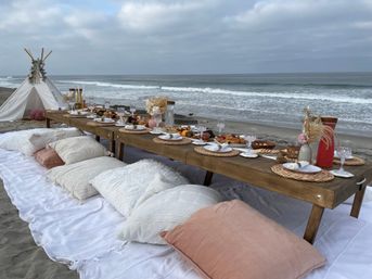 Boho seaside picnic on a sandy beach with a low wooden table, woven placemats, glassware, charcuterie, floral vases, plush cushions and a small teepee by the ocean waves under a cloudy sky.