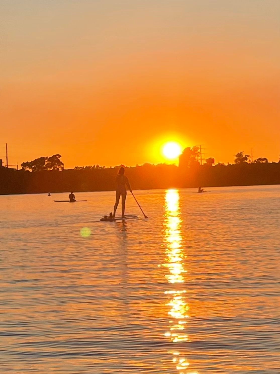 Silhouette of a paddleboarder and distant kayakers gliding on a calm lake at golden sunset, fiery orange sky and a bright sun casting a shimmering reflection across the water