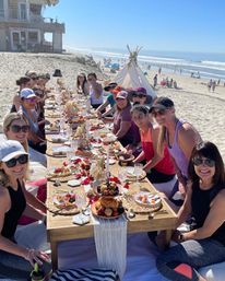 Sunny oceanfront beach brunch: a large group of women seated around a low wooden table on sand, sharing pastries, fruit, crystal glasses and floral decor with a teepee and waves on the horizon.