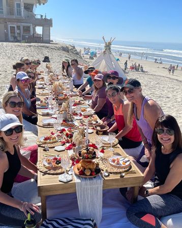 Sunny oceanfront beach brunch: a large group of women seated around a low wooden table on sand, sharing pastries, fruit, crystal glasses and floral decor with a teepee and waves on the horizon.