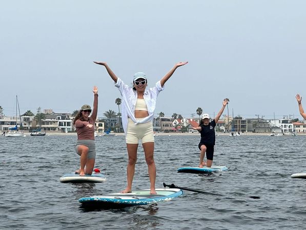 Three paddleboarders striking joyful poses with arms raised on a calm coastal bay, sailboats and waterfront homes visible along the shore under an overcast sky.