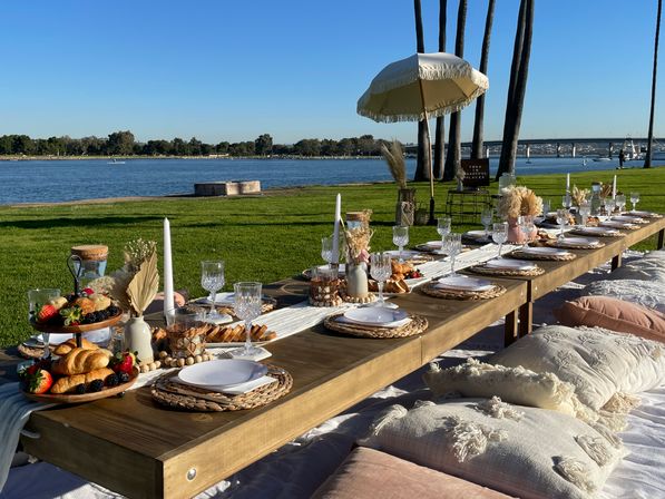 Outdoor waterfront boho picnic on a grassy bay — long low wooden table with woven placemats, white plates, crystal glassware, croissants, berries, candles and pampas grass, surrounded by plush floor cushions and a fringed umbrella with palm trees and a bridge over calm water in the background.