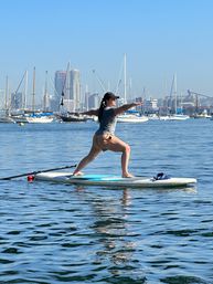 Person doing stand-up paddleboard (SUP) yoga warrior pose on a paddleboard in a marina with anchored sailboats and an urban skyline under a clear blue sky.