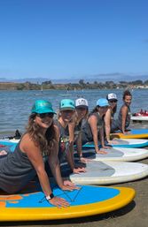 Group of women practicing paddleboard yoga on colorful SUPs at a sunny coastal bay with calm water and distant shoreline