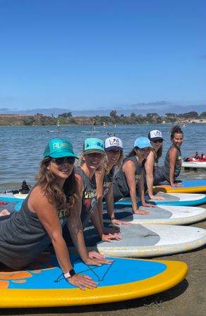 Group of women practicing paddleboard yoga on colorful SUPs at a sunny coastal bay with calm water and distant shoreline