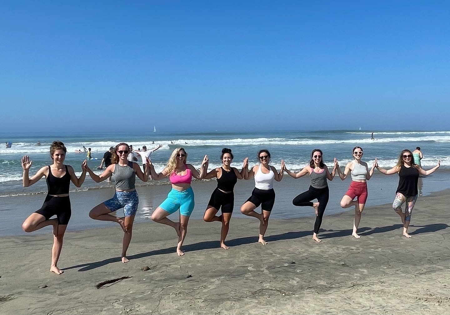 Nine people doing group beach yoga in tree pose, holding hands on a sunny sandy ocean shoreline with gentle waves and a clear blue sky.