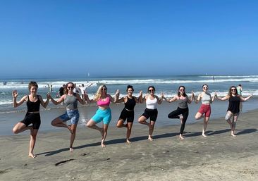 Nine people doing group beach yoga in tree pose, holding hands on a sunny sandy ocean shoreline with gentle waves and a clear blue sky.