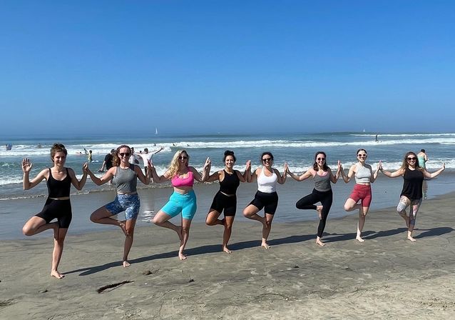 Nine people doing group beach yoga in tree pose, holding hands on a sunny sandy ocean shoreline with gentle waves and a clear blue sky.