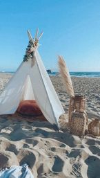 Boho beach teepee on sunlit sand with a rust cushion, wicker lanterns and pampas grass, ocean waves and clear blue sky in the background