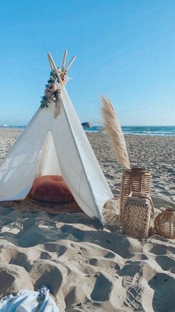Boho beach teepee on sunlit sand with a rust cushion, wicker lanterns and pampas grass, ocean waves and clear blue sky in the background