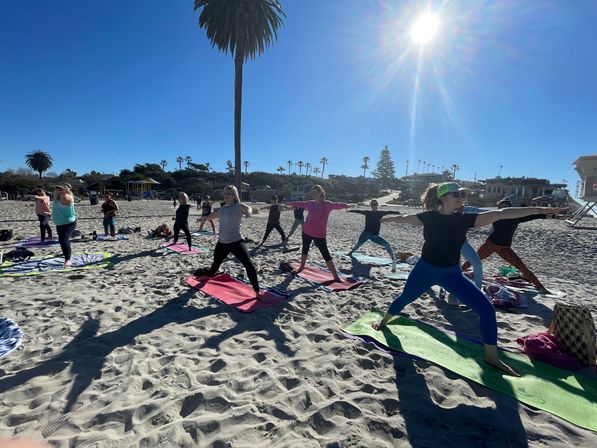 Group doing beach yoga in warrior poses on colorful mats under a bright sun and tall palm trees on a sunny coastal beach