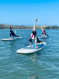 Three paddleboarders practicing SUP yoga on a calm coastal bay under a clear blue sky, kneeling lunge poses on inflatable stand-up paddleboards with shoreline and rolling hills in the background.