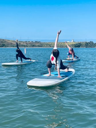 Three paddleboarders practicing SUP yoga on a calm coastal bay under a clear blue sky, kneeling lunge poses on inflatable stand-up paddleboards with shoreline and rolling hills in the background.