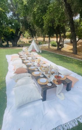 Boho outdoor picnic setup on a green lawn under oak trees — low dark wooden table with wicker chargers, white plates, glassware and candles, blush and cream floor pillows on a large white blanket, small white teepee in the background.