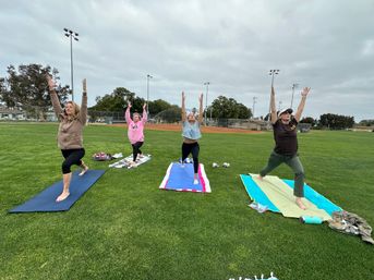 Four people practicing outdoor yoga on colorful mats on a grassy baseball field in a local park, arms raised toward an overcast sky.