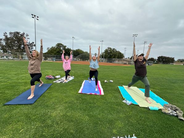 Four people practicing outdoor yoga on colorful mats on a grassy baseball field in a local park, arms raised toward an overcast sky.