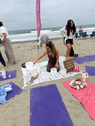 Oceanfront beach scene: woman arranging crystal singing bowls on a blanket for a beachfront sound bath birthday, balloons and a birthday sign nearby, purple and pink yoga mats on the sand with waves in the background.