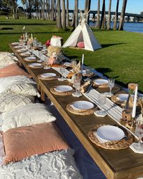 Boho-chic waterfront picnic in a grassy park — long low wooden table set with wicker chargers, white plates, crystal glassware, candles and dried floral arrangements, surrounded by plush pillows, a small white teepee, palm trees and a river bridge in the background.