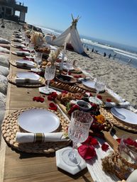 Boho beach picnic on a sunny coastline — long low wooden table on sand set with woven placemats, white plates, crystal goblets, gold‑banded napkins, charcuterie boards, rose petals and a small floral teepee with ocean waves in the background.