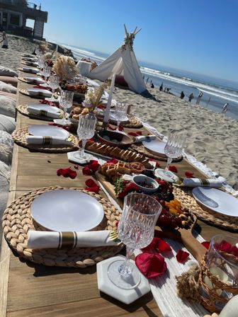 Boho beach picnic on a sunny coastline — long low wooden table on sand set with woven placemats, white plates, crystal goblets, gold‑banded napkins, charcuterie boards, rose petals and a small floral teepee with ocean waves in the background.