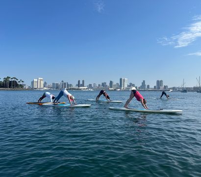 Group practicing paddleboard yoga on San Diego Bay — downward dog on SUPs with calm blue water, downtown skyline and palm trees under a clear blue sky