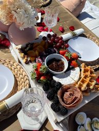 Sunlit outdoor picnic-style charcuterie board on a wooden table with brie, waffles, salami rose, strawberries, blackberries, red grapes, dried apricots, mixed nuts and fig jam, crystal glasses, woven placemats and a blush floral vase.