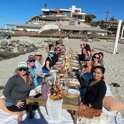 Sunny beach brunch: a long low wooden table on white blankets with a group of people sharing pastries, drinks and tableware on the sand, coastal homes and blue sky in the background.
