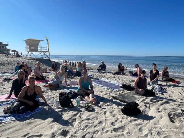Sunlit beach yoga group on sandy shore with a lifeguard tower and calm ocean under a clear blue sky.