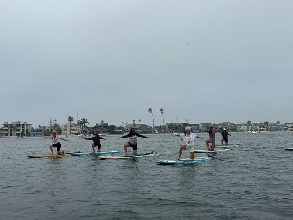 Group practicing paddleboard yoga on a calm coastal bay, kneeling on SUP boards with arms outstretched in front of beachfront homes and palm trees under an overcast sky.