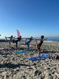 Four people balancing in dancer (standing bow) pose on yoga mats at a sunny sandy beach by the ocean under a clear blue sky — seaside group yoga session.