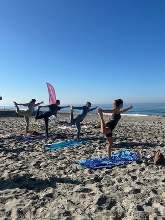 Four people balancing in dancer (standing bow) pose on yoga mats at a sunny sandy beach by the ocean under a clear blue sky — seaside group yoga session.
