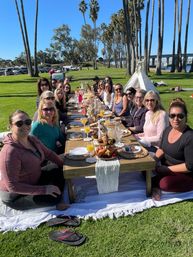 Sunny waterfront brunch picnic: long low wooden table with pastries, mimosas and candles, a group of adults gathered on a grassy coastal park lined with tall palm trees and a bridge in the background.