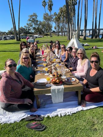 Sunny waterfront brunch picnic: long low wooden table with pastries, mimosas and candles, a group of adults gathered on a grassy coastal park lined with tall palm trees and a bridge in the background.
