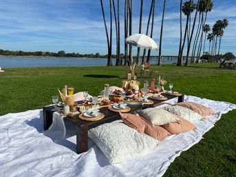Sunny boho picnic at a palm-lined waterfront park: low wooden table on a white blanket with cushions, wicker chargers, glassware, charcuterie boards and a fringed umbrella under a blue sky.