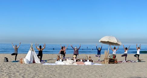 Beach yoga group doing tree pose on a sunny sandy oceanfront shore, facing calm blue waves with a cozy picnic spread, teepee and fringed umbrella — seaside wellness gathering