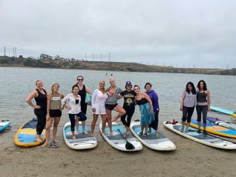 Smiling group of women posing on stand-up paddleboards on a sandy shore beside calm water under an overcast sky, with rolling hills and power lines in the background.