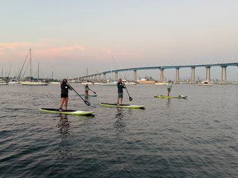 Four paddleboarders on calm San Diego Bay at dusk with sailboats nearby and the Coronado Bridge in the background.