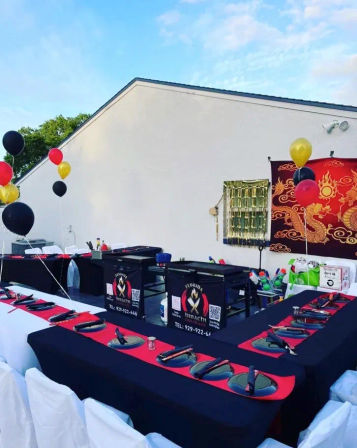 Outdoor hibachi-style backyard setup with U-shaped black-and-red tables, place settings and chopsticks, portable teppanyaki grills, red-and-gold dragon backdrop and black, red and gold balloons.