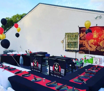 Festive outdoor hibachi-style patio setup with U-shaped black-and-red table linens, portable teppanyaki grills, white-covered chairs, dragon tapestry backdrop and red, black and gold balloons.