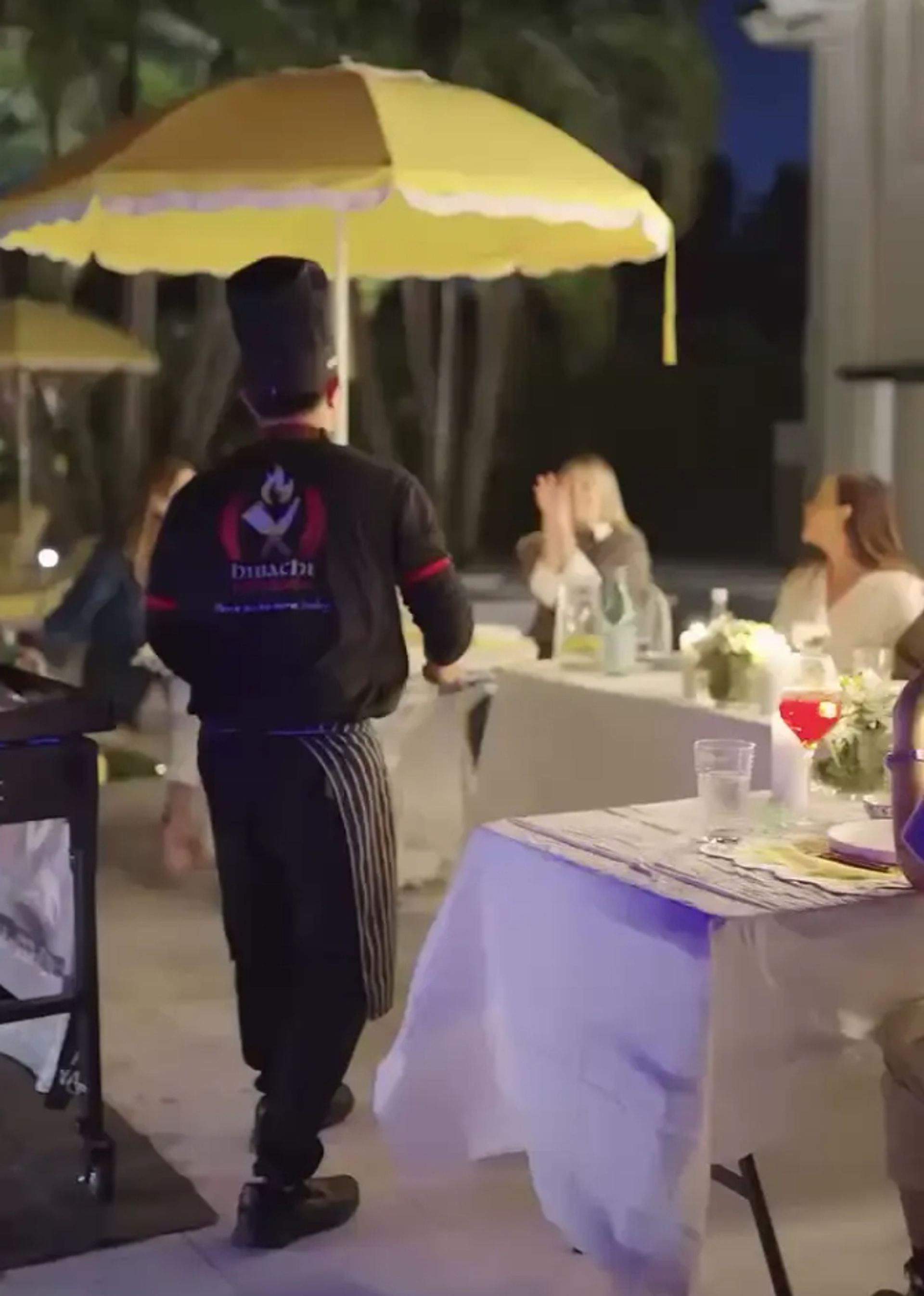Evening outdoor patio dining scene with a chef in a black uniform walking under a vibrant yellow umbrella toward guests seated at white-clothed tables with cocktails and soft ambient lighting.