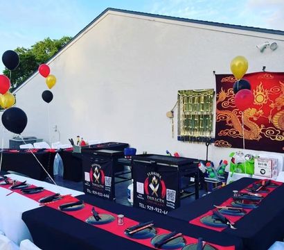 Outdoor hibachi-style party setup on a patio: U-shaped black-and-red tables with white chairs, place settings with chopsticks and round plates, central portable grill stations, red/gold/black balloons and a decorative red dragon backdrop.