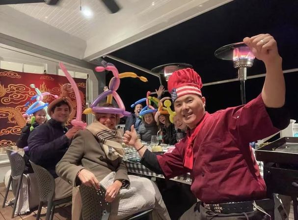 Cheerful group wearing colorful balloon hats at an outdoor nighttime patio dinner, a smiling chef in red giving thumbs-up beside a checkered table with glowing patio heaters.