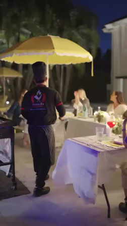 Outdoor evening dinner scene with a waiter in a chef hat and striped apron walking beneath a bright yellow patio umbrella toward a candlelit table of seated guests with drinks and floral centerpieces.